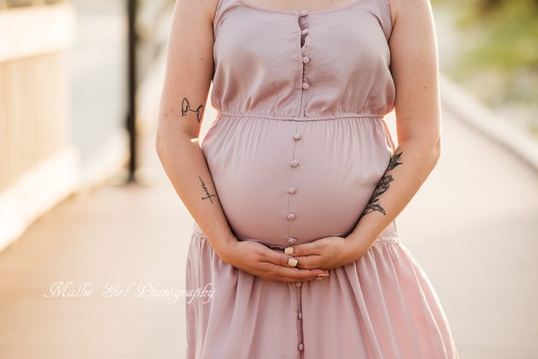 Pregnant Woman in a Soft Pink Dress Stands Outdoors, Hands Gently Cradling Her Baby Bump, with Visible Arm Tattoos.