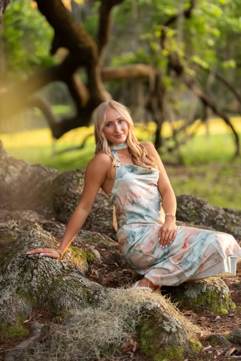 Blonde Woman in a Pastel Floral Dress Sits on Mossy Rocks in a Sunlit Forest Clearing, Smiling at the Camera.
