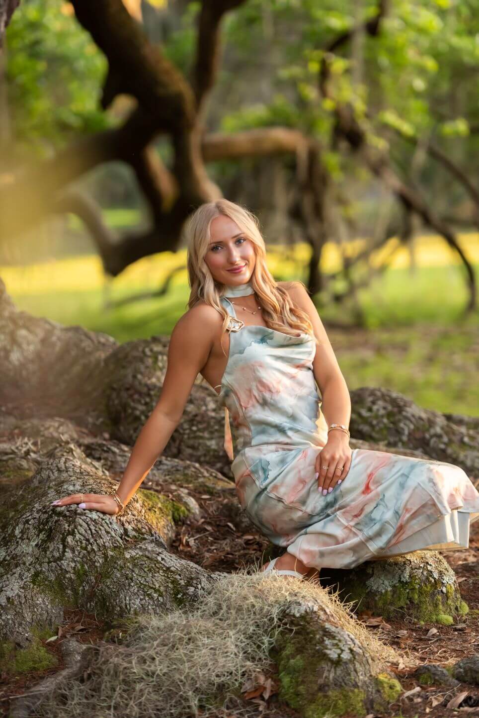 Blonde Woman in a Pastel Floral Dress Sits on Mossy Rocks in a Sunlit Forest Clearing, Smiling at the Camera.